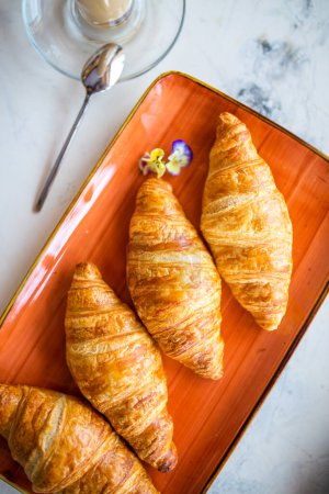 Photo for A delightful breakfast with croissants and coffee on a marble table top view. Croissants are golden brown on an orange plate, coffee with froth. - Royalty Free Image