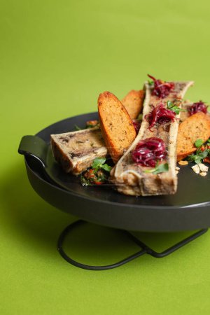 Gebratenes Knochenmark mit buttrigem Reichtum auf knusprigem Brot, garniert mit frischen Kräutern, serviert auf einem schwarzen Teller vor grünem Hintergrund.