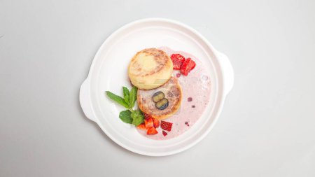 Foto de Tortitas redondas y esponjosas de requesón rematadas con fresas vibrantes y arándanos en un plato blanco, perfectas para un delicioso desayuno. - Imagen libre de derechos