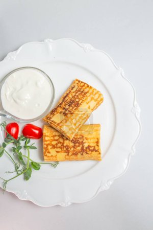 Delicious blintzes topped with creamy sour cream, served with fresh cherry tomatoes on a elegant white plate.