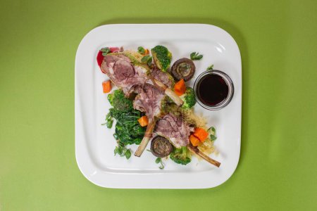 Téléchargez les photos : Vue du dessus d'un plat avec côtes d'agneau juteuses, légumes cuits au four et sauce rouge parfumée sur fond vert - en image libre de droit