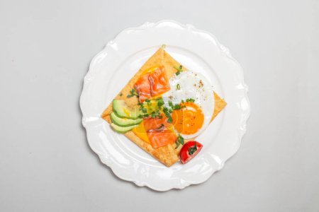 Nahaufnahme eines Crêpes mit Räucherlachs, Avocado, Ei und Tomaten auf einem weißen Teller. Quadratische Form, elegante Präsentation.