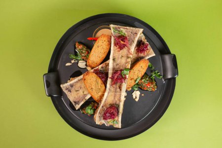 Beef bone marrow with toasts and herbs on a black plate, set on a vibrant green background, a delectable and elegant dish.