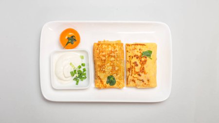 Two folded stuffed savory crepes with sour cream and chives garnish on a white plate on a white background.