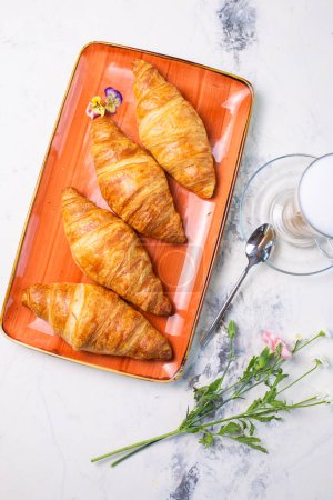 Un delicioso desayuno con cruasanes y café con vistas a la mesa de mármol. Las croissants son de color marrón dorado en un plato naranja, café con espuma.