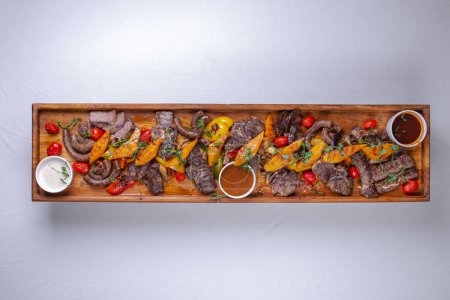 A long wooden platter filled with an assortment of meats and vegetables, including steak, sausage, chicken, potatoes, carrots, and peppers.
