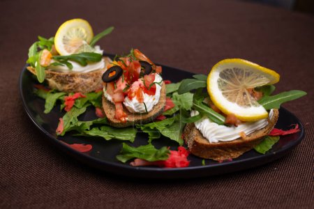 A variety of bruschettas with cream cheese, tomatoes, olives, lemon on a black plate, with lettuce and red pickled ginger garnish