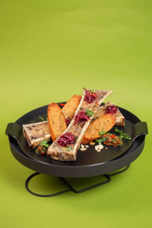 Roasted bone marrow with buttery richness on crusty bread, garnished with fresh herbs, served on a black plate against a green backdrop.