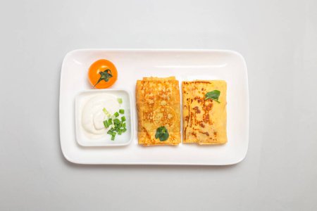 Dos crepes rellenos rellenos salados con crema agria y cebollino adornan en un plato blanco sobre un fondo blanco.