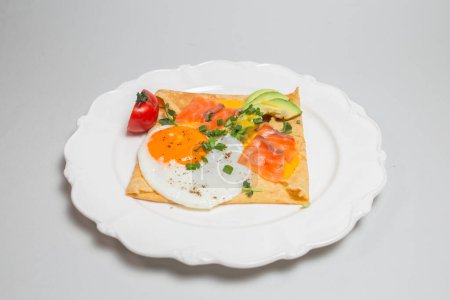 Close-up shot of a crepe with smoked salmon, avocado, egg, and tomatoes on a white plate. Square shape, elegant presentation.