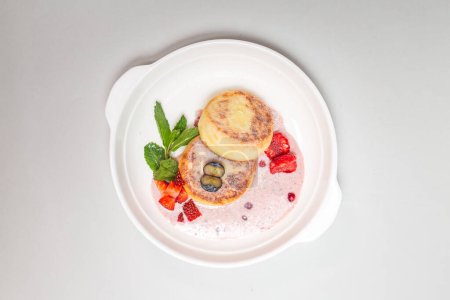 Foto de Tortitas redondas y esponjosas de requesón rematadas con fresas vibrantes y arándanos en un plato blanco, perfectas para un delicioso desayuno. - Imagen libre de derechos