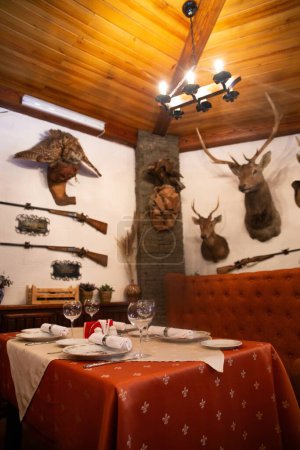 Comedor rústico con mesa de madera, mantel rojo y decoración de trofeos que incluye astas, pistolas y un búho montado.