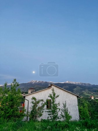 A house in mountains at night. Full moon, bright stars, white exterior, grey roof. Surrounded by trees and mountains.