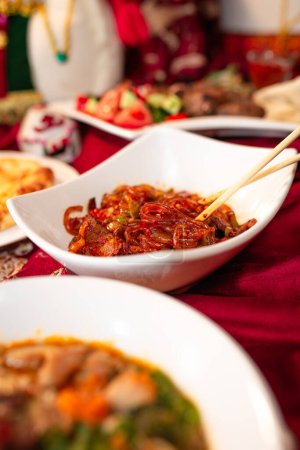 Ein Teller mit leckeren Nudeln mit Fleisch und Gemüse sitzt auf einer roten Tischdecke, dazu andere Gerichte und ein Getränk im Hintergrund..
