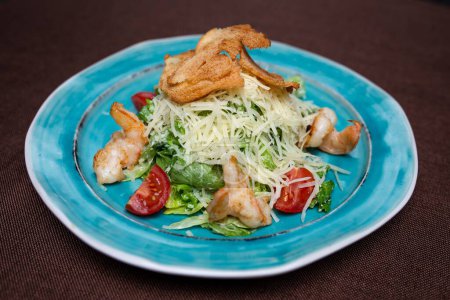 Healthy and delicious caesar salad with fresh greens, juicy tomatoes, and tasty shrimps on a blue plate. Isolated on a brown background.