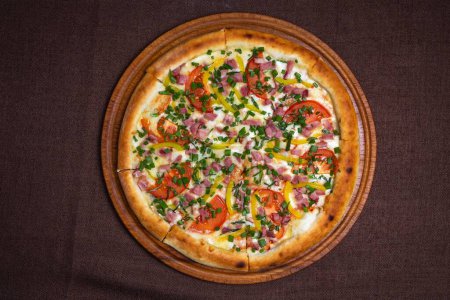 Leckere Pizza mit Schinken, Tomaten, Paprika und frischen Kräutern auf einem Holzbrett. Vereinzelt auf braunem Hintergrund.