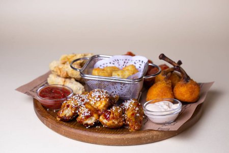 Photo for A variety of fried chicken wings, potato balls, and cheese sticks with dipping sauces on a wooden platter. - Royalty Free Image