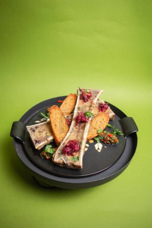 Beef bone marrow with toasts and herbs on a black plate, set on a vibrant green background, a delectable and elegant dish.