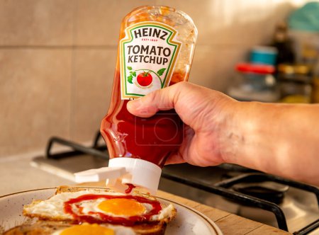 Foto de London. UK- 01.22.2023. A person squeezing a bottle of Heinz tomato ketchup on to fried eggs on toast. - Imagen libre de derechos