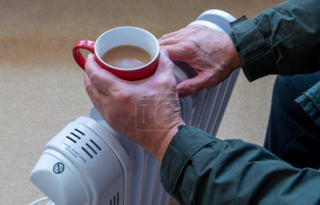 Eine Person hält eine heiße Tasse Tee in der Hand und wärmt die Hände auf einem tragbaren, ölgefüllten Elektroheizer in einem Wohnzimmer.