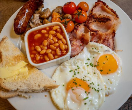 Primer plano de un plato completo de desayuno inglés con huevos, tocino, salchichas, frijoles horneados, champiñones, tomates cherry y pan con mantequilla.