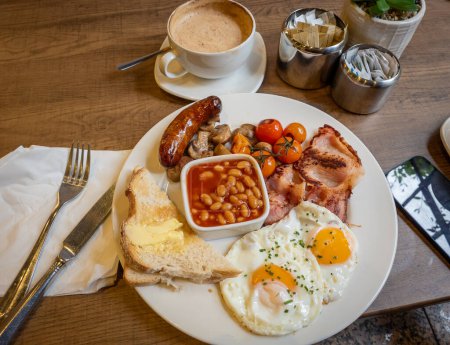 A plate of full English breakfast served on a table with a cup of coffee.