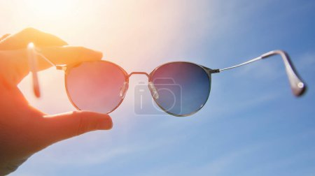 Téléchargez les photos : Main tenant des lunettes de soleil rondes élégantes sur un ciel lumineux et un fond de soleil. Porter des lunettes de soleil par une journée ensoleillée d'été. Un homme regarde le soleil brillant à travers des lunettes de soleil polarisées. Humeur estivale - en image libre de droit