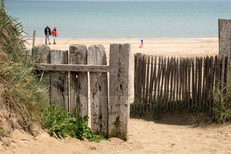 Utah Beach in Normandy, Two unrecognizable people. USA Flag on wood sea fence, Grass and sand. Sunny sky light ble clouds. High quality photo