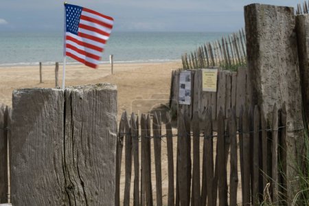 Utah Beach in Normandy, France. Mid range USA flag on wood sea fence, grass and sand dunes. Sunny sky light ble clouds. High quality photo