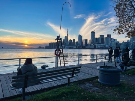 Foto de Toronto, canada - 24 October 2022: people sitting on bench at waterfront and overlooking sunset and skyline of Toronto over lake ontario - Imagen libre de derechos