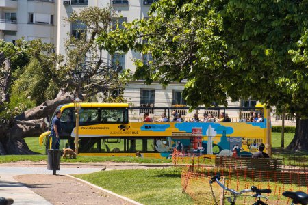 Foto de Buenos aires, Argentina - 27 de octubre de 2022: un tour amarillo hop on hop de bus turístico pasa por el parque Recoleta donde un hombre pasea a un perro - Imagen libre de derechos