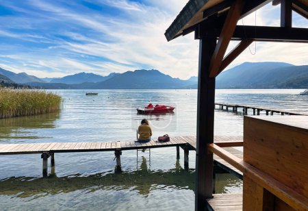 Photo for Annecy, France - October 15 2021 : a woman is sitting alone on a calm day with almost blue sky on a wooden pier or board walk overlooking the lake - Royalty Free Image
