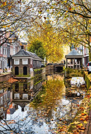 Photo for Gouda, Netherlands - November 11 2020 : a typical idyllic view of a dutch historic town with old houses along a canal with trees and reflection and fall leafs floating on the water - Royalty Free Image