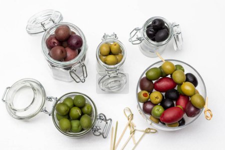 Foto de Olives in glass jars. Skewers on the table. Flat lay. White background. - Imagen libre de derechos