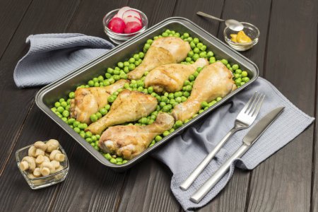 Chicken drumsticks and green peas in metal baking tray. Mustard, radish on table. Fork and knife on gray napkin. Dark wooden background.