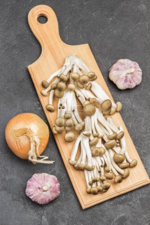 Broun Shimeji mushrooms on cutting board. Onions and garlic on table. Black background