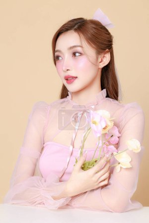 Téléchargez les photos : Jeune femme asiatique mignonne dans une robe élégante rose, maquillage de style coréen, hydratée, lisse, peau parfaite avec des fleurs sur un fond beige. Traitement du visage, cosmétologie, chirurgie plastique. - en image libre de droit