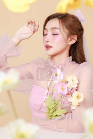 Photo for Young cute Asian woman in a pink elegant dress, Korean style makeup, moisturized, smooth, perfect skin with flowers on a beige background. Facial treatment, Cosmetology, plastic surgery. - Royalty Free Image