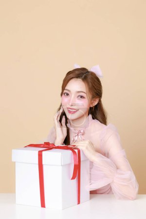 Foto de Mujer asiática linda joven feliz en un vestido elegante rosa, maquillaje de estilo coreano, piel hidratada, lisa, perfecta con caja de regalo sobre un fondo beige. Tratamiento facial, Cosmetología, cirugía plástica. - Imagen libre de derechos
