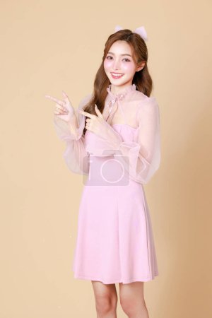 Téléchargez les photos : Jeune femme asiatique mignonne dans une robe élégante rose, maquillage de style coréen, hydratée, lisse, peau parfaite pointant vers copier l'espace sur un fond beige. Traitement du visage, cosmétologie, chirurgie plastique. - en image libre de droit