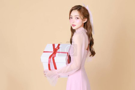 Photo for Young cute Asian woman happy in a pink elegant dress, Korean style makeup, moisturized, smooth, perfect skin with Gift box on a beige background. Facial treatment, Cosmetology, plastic surgery. - Royalty Free Image