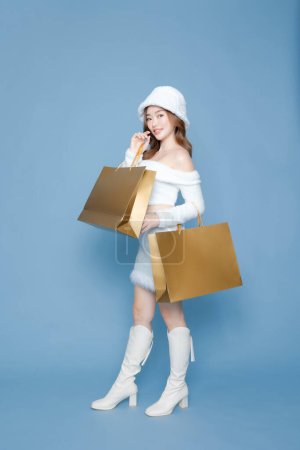 Photo for Asian happy woman with perfect body posing in studio. Luxury fashionable female model in white dress with golden shopping bags on blue isolated background. E-commerce, Online shopping Concept. - Royalty Free Image