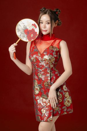 Photo for Beautiful young Asian woman with clean fresh skin wearing traditional cheongsam qipao dress holding fan posing on red background. Portrait of female model in studio. Happy Chinese new year. - Royalty Free Image