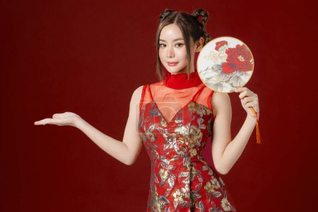 Téléchargez les photos : Belle jeune femme asiatique avec une peau propre et fraîche portant la robe traditionnelle cheongsam qipao tenant ventilateur posant sur fond rouge. Portrait de mannequin femme en studio. Bonne année chinoise. - en image libre de droit