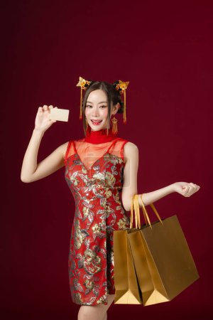 Beautiful Asian woman with clean fresh skin wearing traditional cheongsam dress holding credit card Gold shopping bags on red background. Happy Chinese new year.