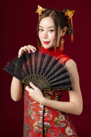 Téléchargez les photos : Belle jeune femme asiatique avec une peau propre et fraîche portant la robe traditionnelle cheongsam qipao tenant ventilateur posant sur fond rouge. Portrait de mannequin femme en studio. Bonne année chinoise. - en image libre de droit