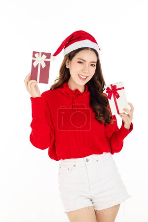 Photo for Cheerful attractive Asian woman wearing a red dress with a Santa hat is holding a gift box on white isolated background. Beautiful lady with long black hair. - Royalty Free Image