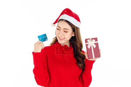 Foto de Alegre atractiva mujer asiática con un vestido rojo con un sombrero de Santa es la celebración de una caja de regalo y tarjeta de crédito sobre fondo blanco aislado. Hermosa dama con el pelo largo y negro. - Imagen libre de derechos