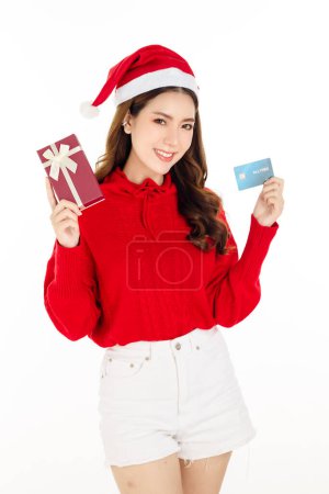 Téléchargez les photos : Joyeux attrayant asiatique femme portant une robe rouge avec un chapeau Santa tient une boîte-cadeau et carte de crédit sur fond blanc isolé. Belle dame aux longs cheveux noirs. - en image libre de droit