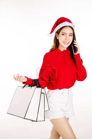 Téléchargez les photos : Joyeux attrayant asiatique femme portant une robe rouge avec un chapeau de Père Noël tient un sac à provisions sur fond blanc isolé. Belle dame aux longs cheveux noirs utilise le téléphone portable shopping en ligne. - en image libre de droit